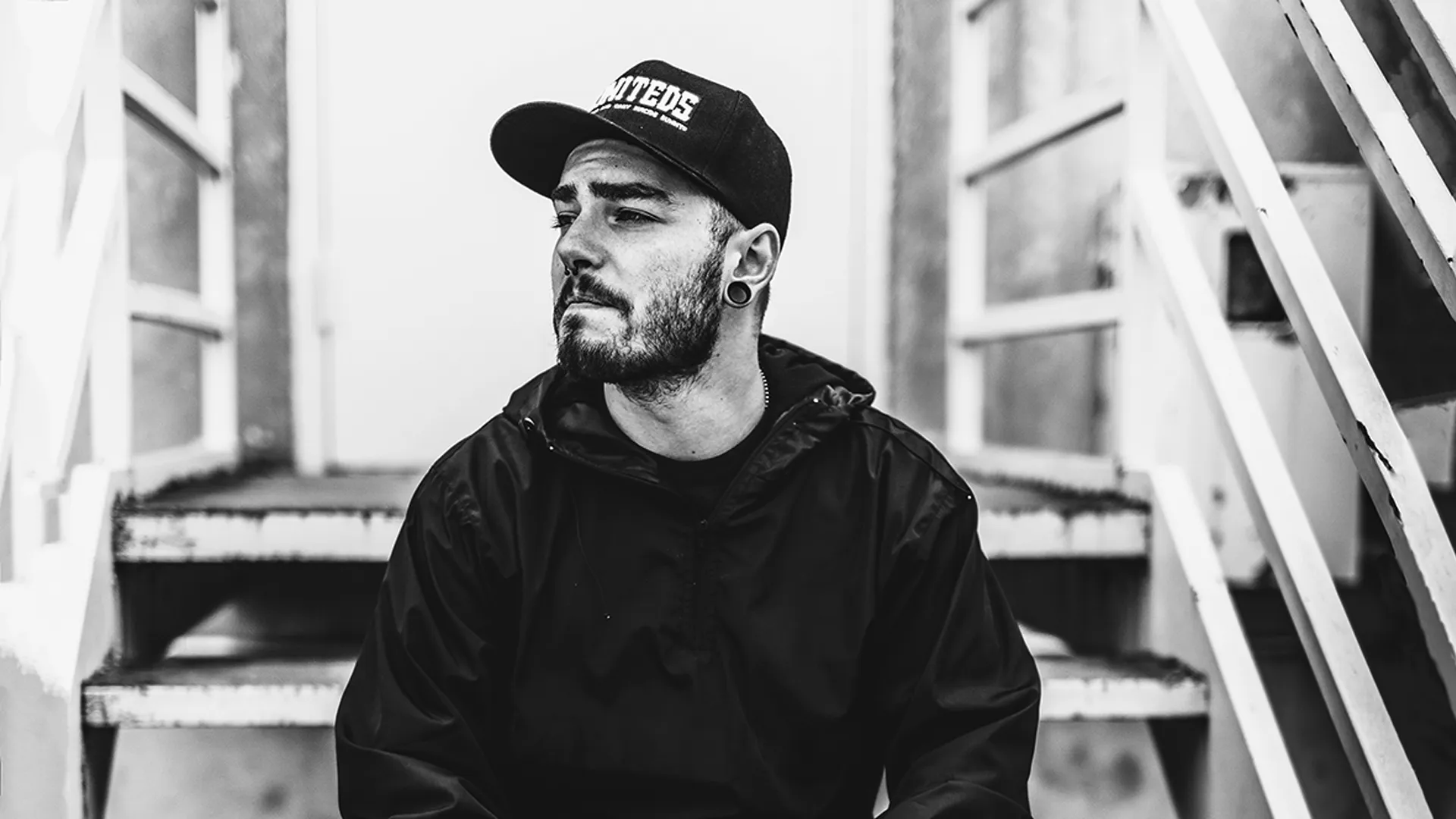 Black and white photo of a bearded man (James Alexzander) wearing a cap and a jacket, with a contemplative expression, standing in front of industrial stairs.